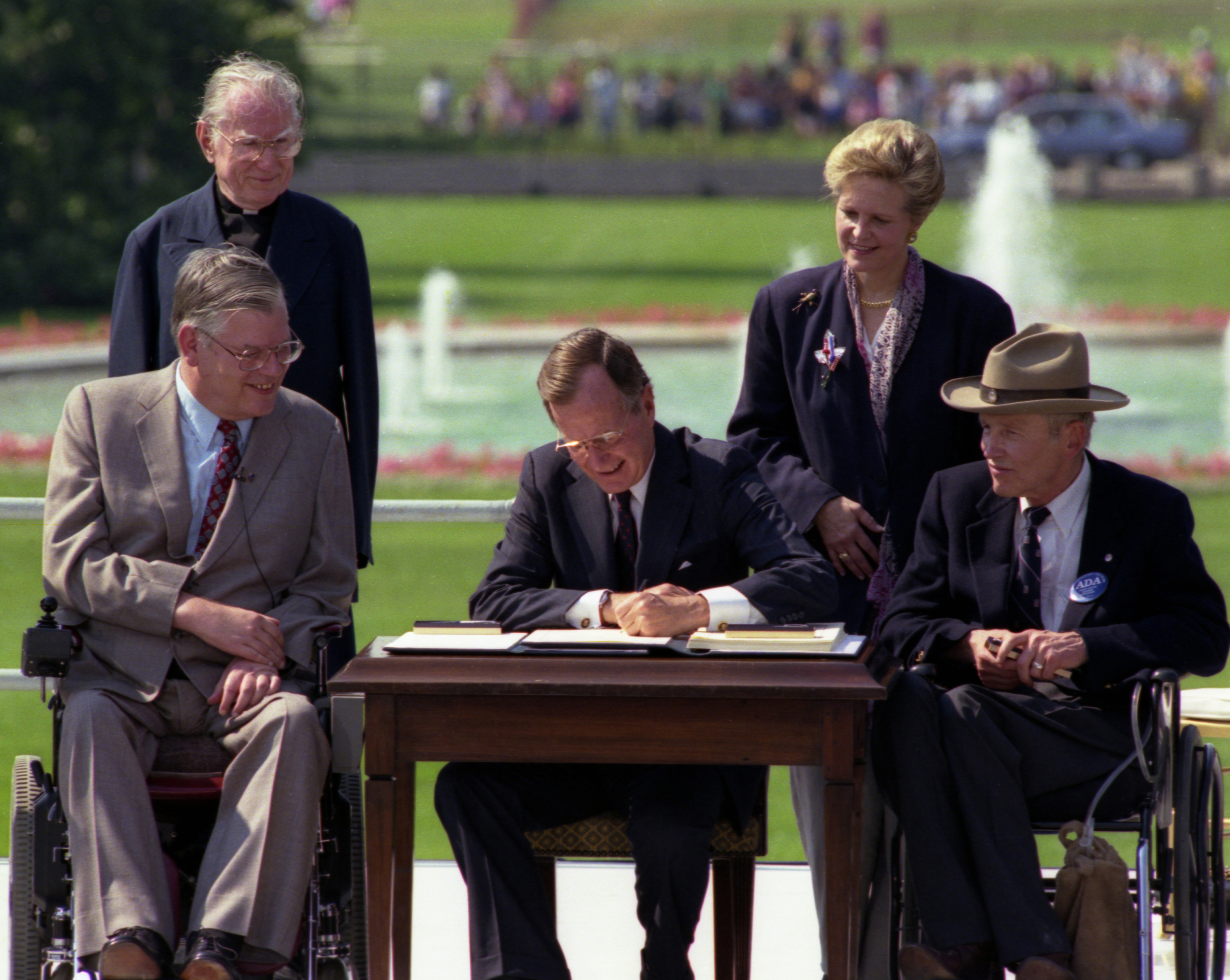 2 people in wheelchairs and 2 standing people look on as a man signs a document.